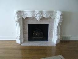 White marble fireplace with ornate carvings and black firebox, set against a white wall and wood floor.