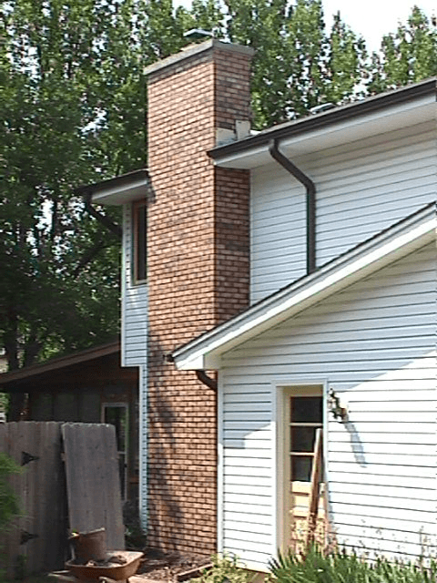Brick chimney attached to a white house with dark gutters, trees in the background.