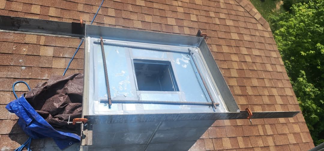 A metal skylight frame is set on a brown shingle roof, with a small tarp and debris nearby.