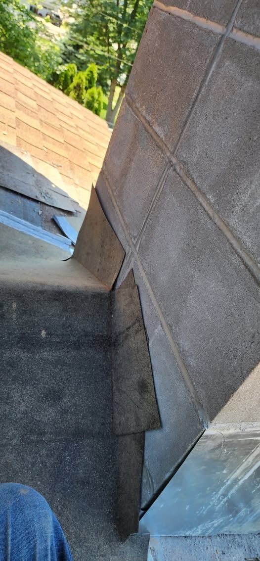 Close-up of a roof corner where two sections meet. Flashing and shingles are visible; sky and trees in the background.