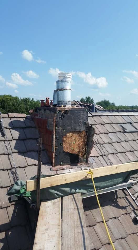 Chimney on a roof: crumbling brick structure with metal flue, viewed against a blue sky.