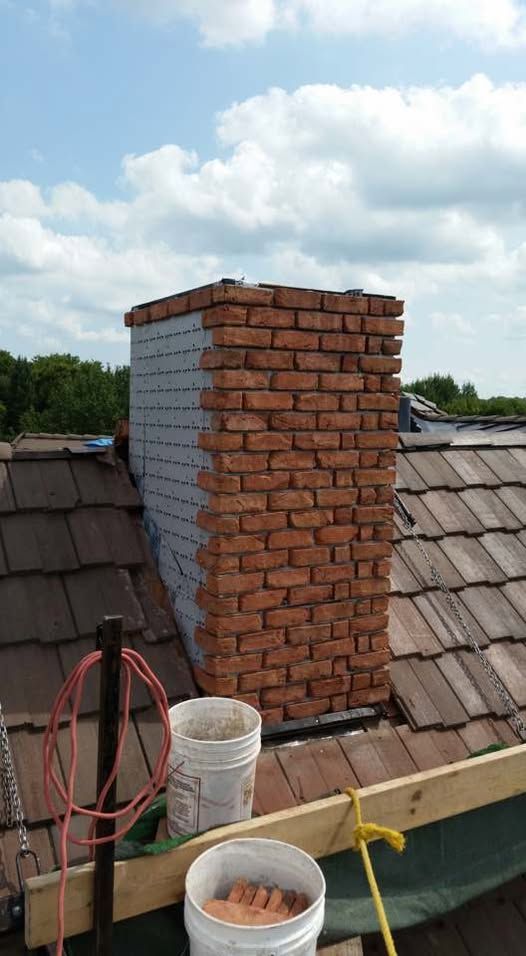 Chimney under construction on a rooftop with red bricks and grey siding. Buckets and tools are visible.