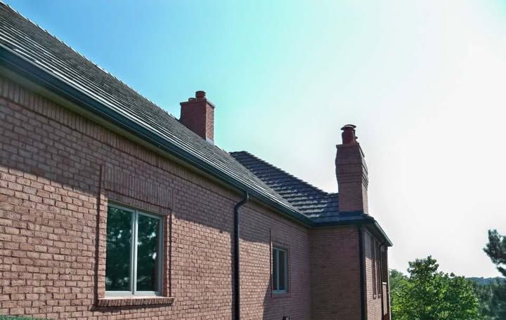 Red brick building with gray roof, black gutters, two chimneys, and two visible windows under a blue sky.
