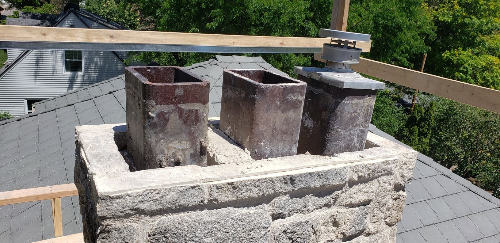 Chimney on a roof, with three flues and wooden supports above, green foliage in the background.