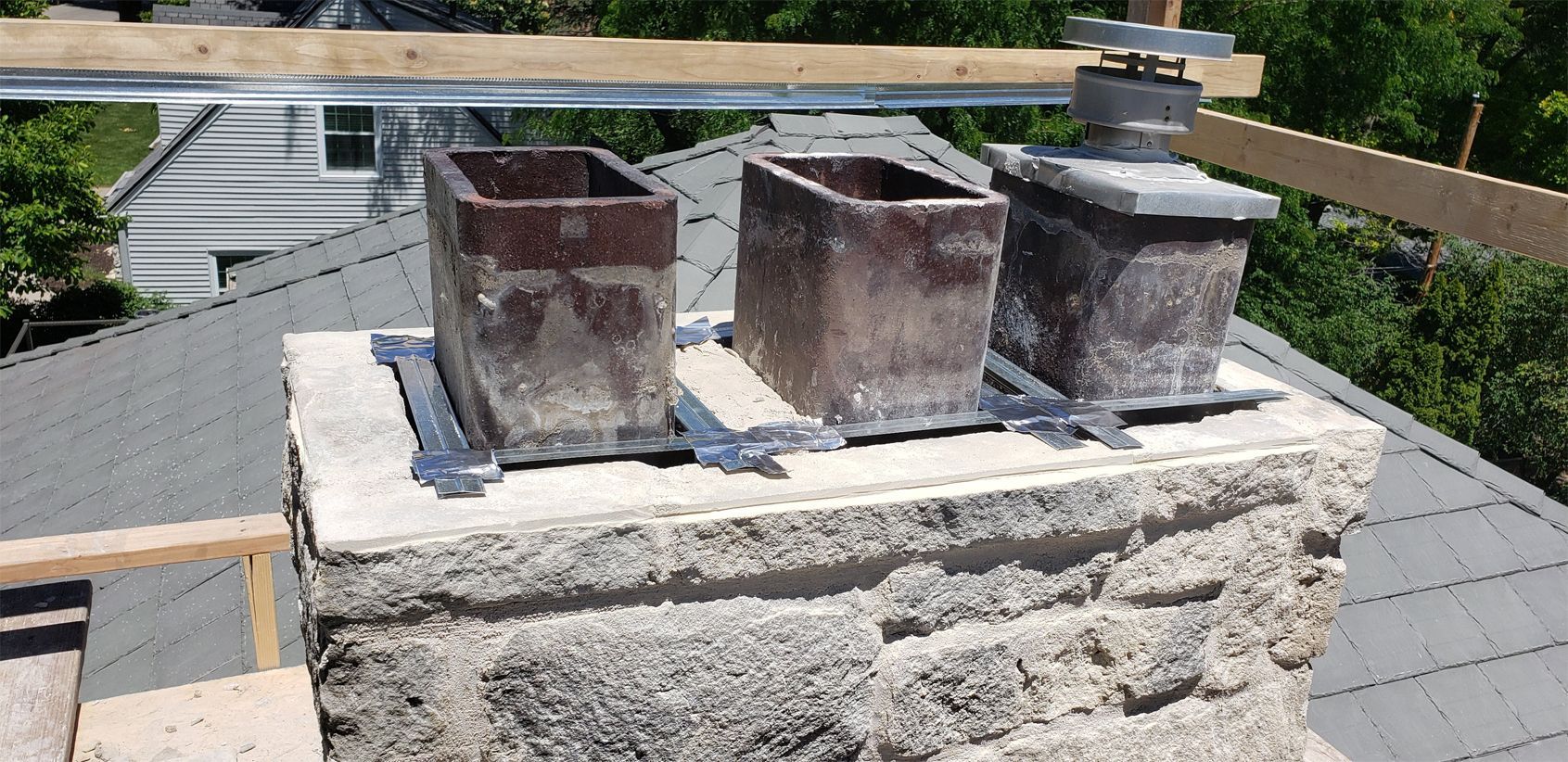 Chimney with three flues on a gray tiled roof. Wooden scaffolding and green foliage are visible in the background.