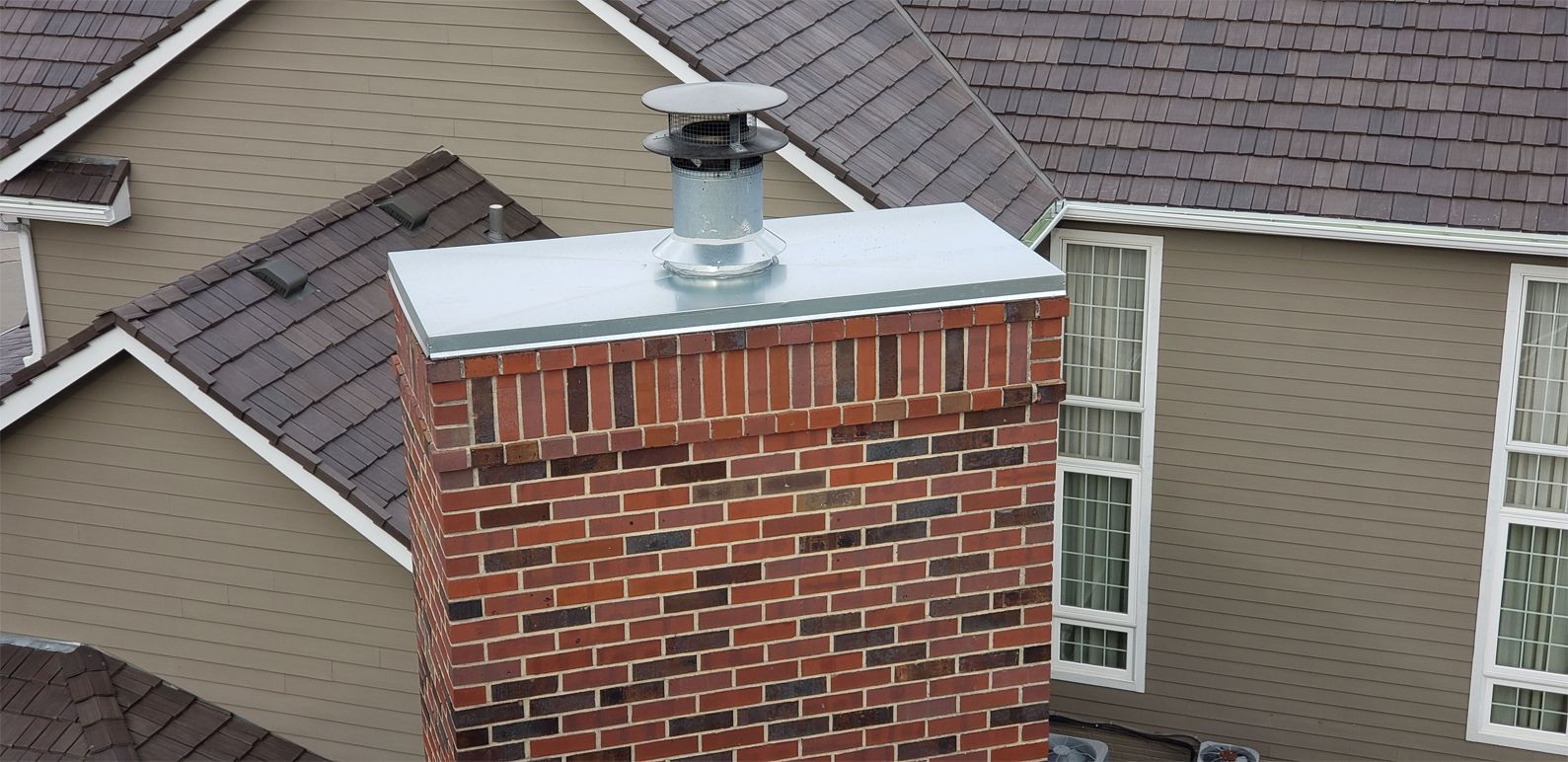 Brick chimney with a metal cap and chimney cap, on a brown roof.