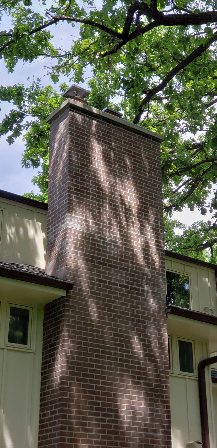 Tall brick chimney on a house with sunlight dappling the surface, trees in the background.