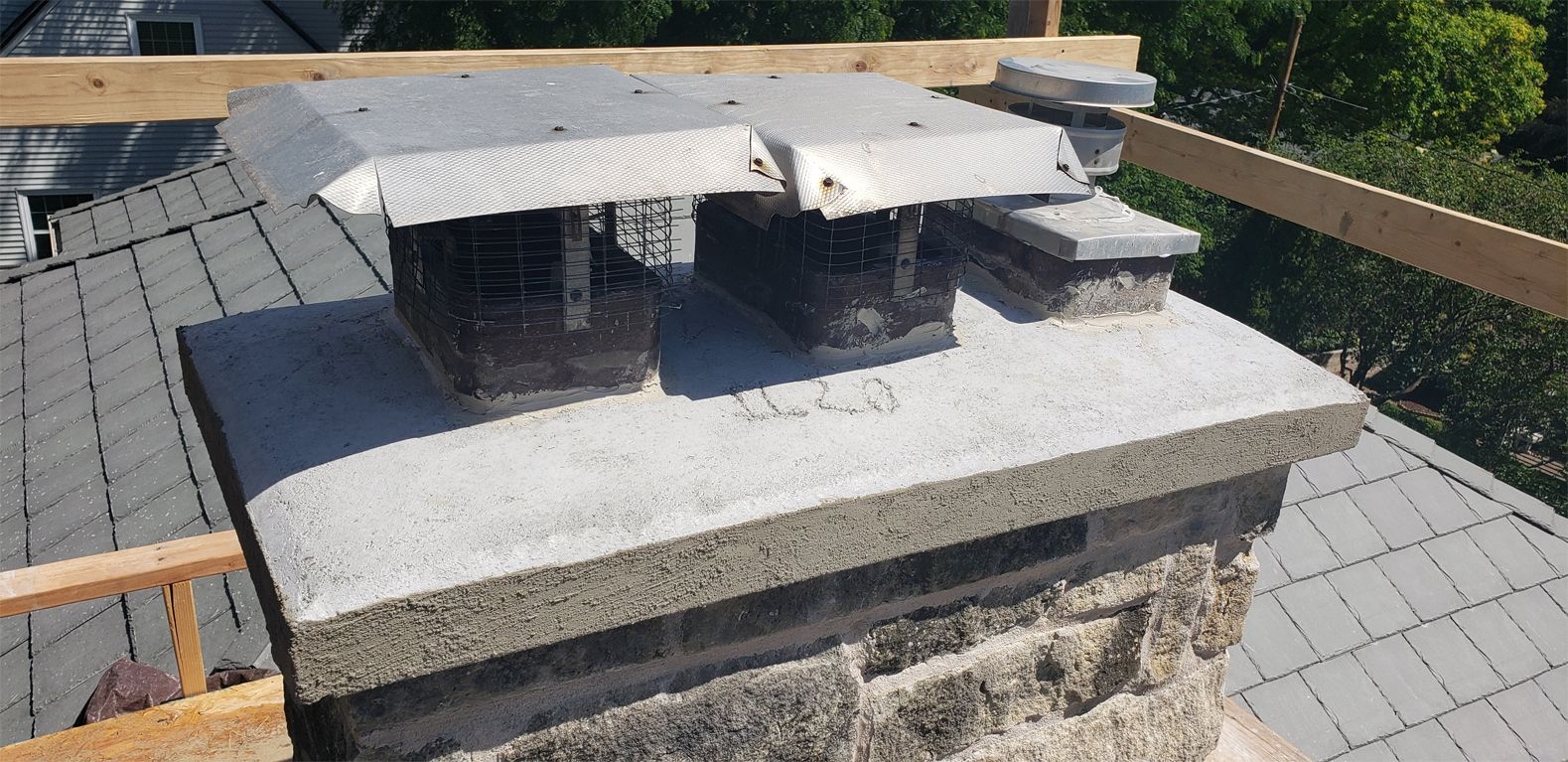 Chimney on a gray roof, with metal chimney caps and a wooden beam above.