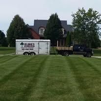A truck and trailer are parked in a lush green field in front of a house.