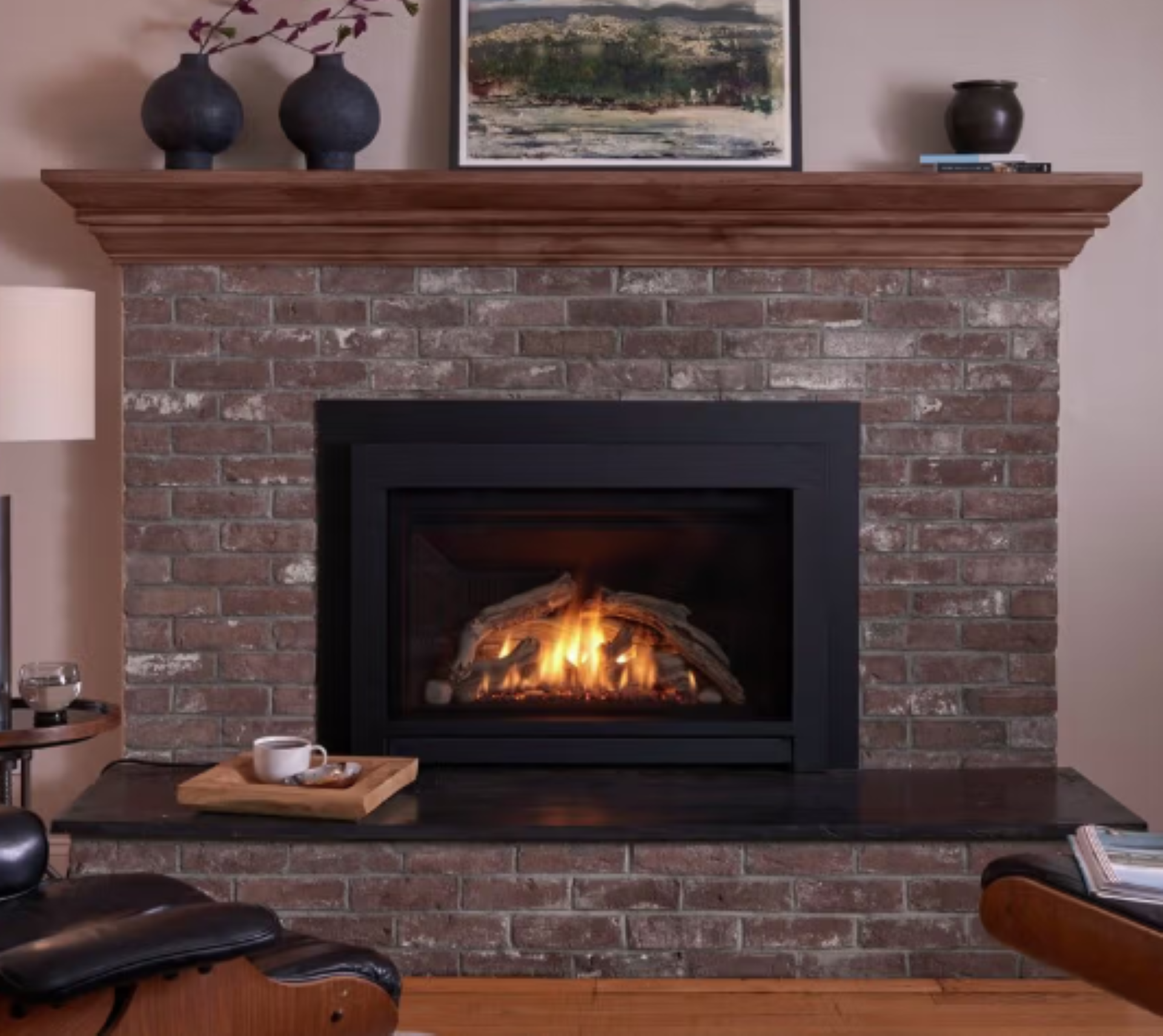 Brick fireplace with a lit fire, wooden mantel, and framed landscape print in a cozy living room.