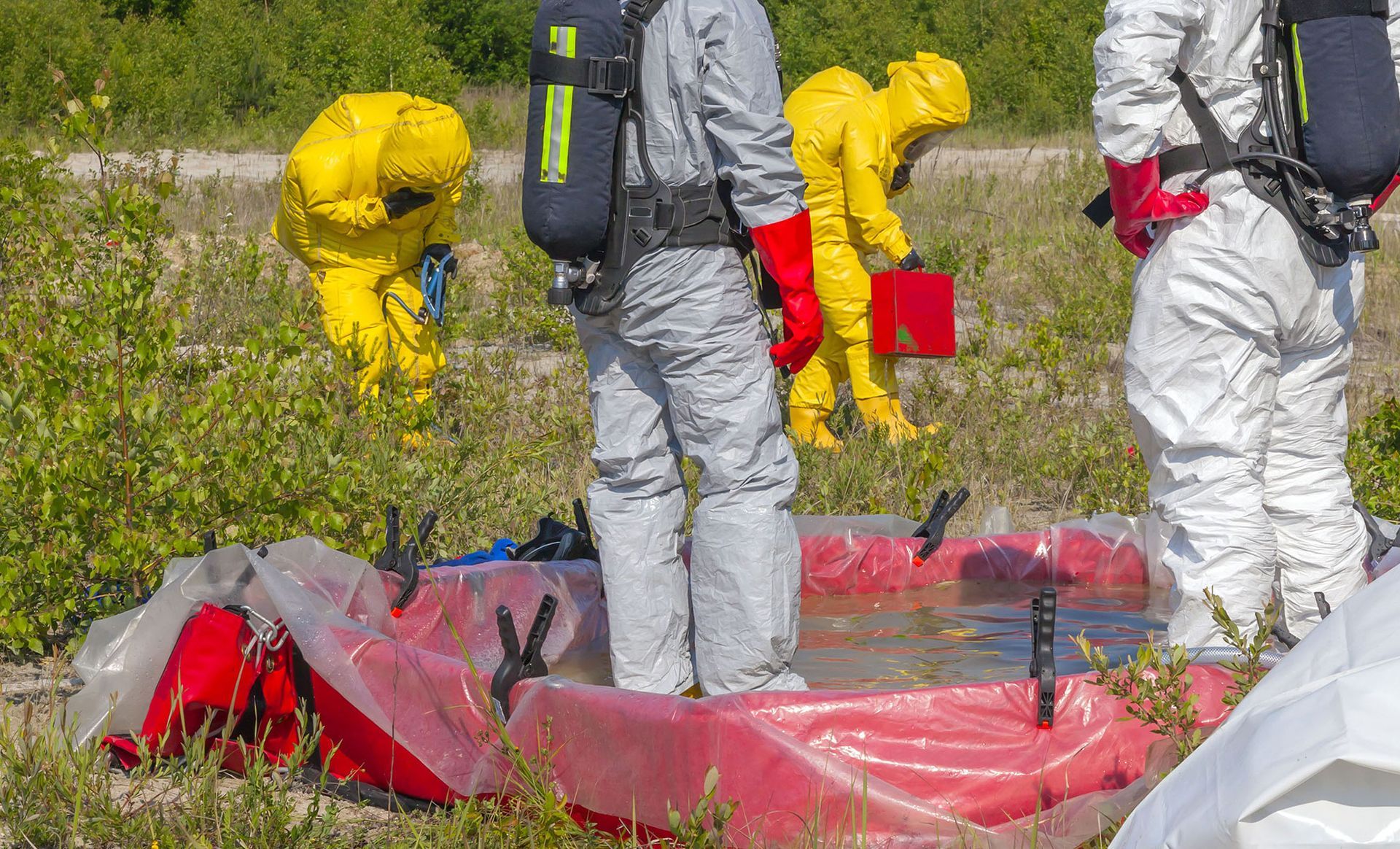 People in hazmat suits working in a grassy area near a red containment pool.