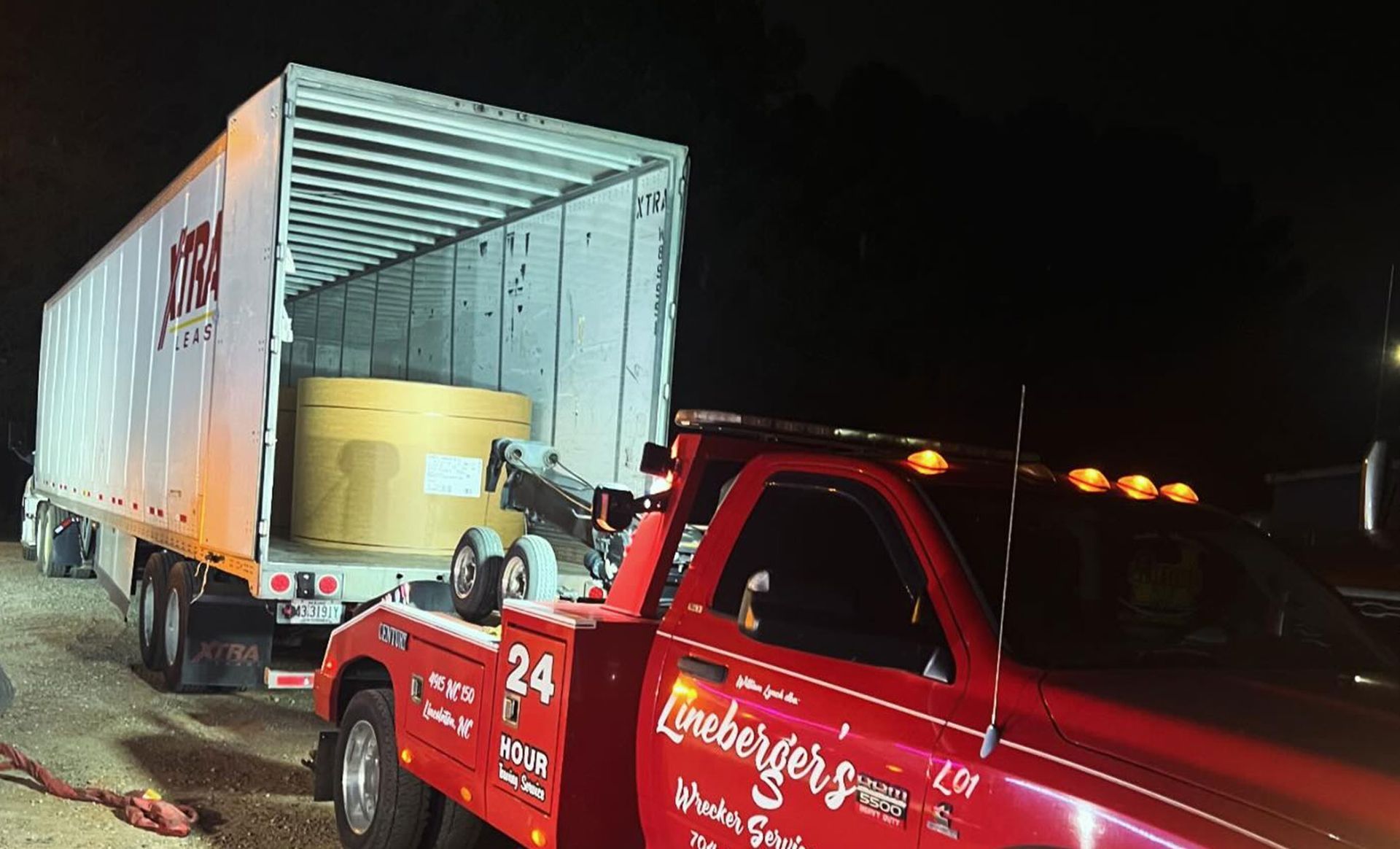 A red tow truck at night hooking up to a semi-trailer. The trailer contains a large, cylindrical object.