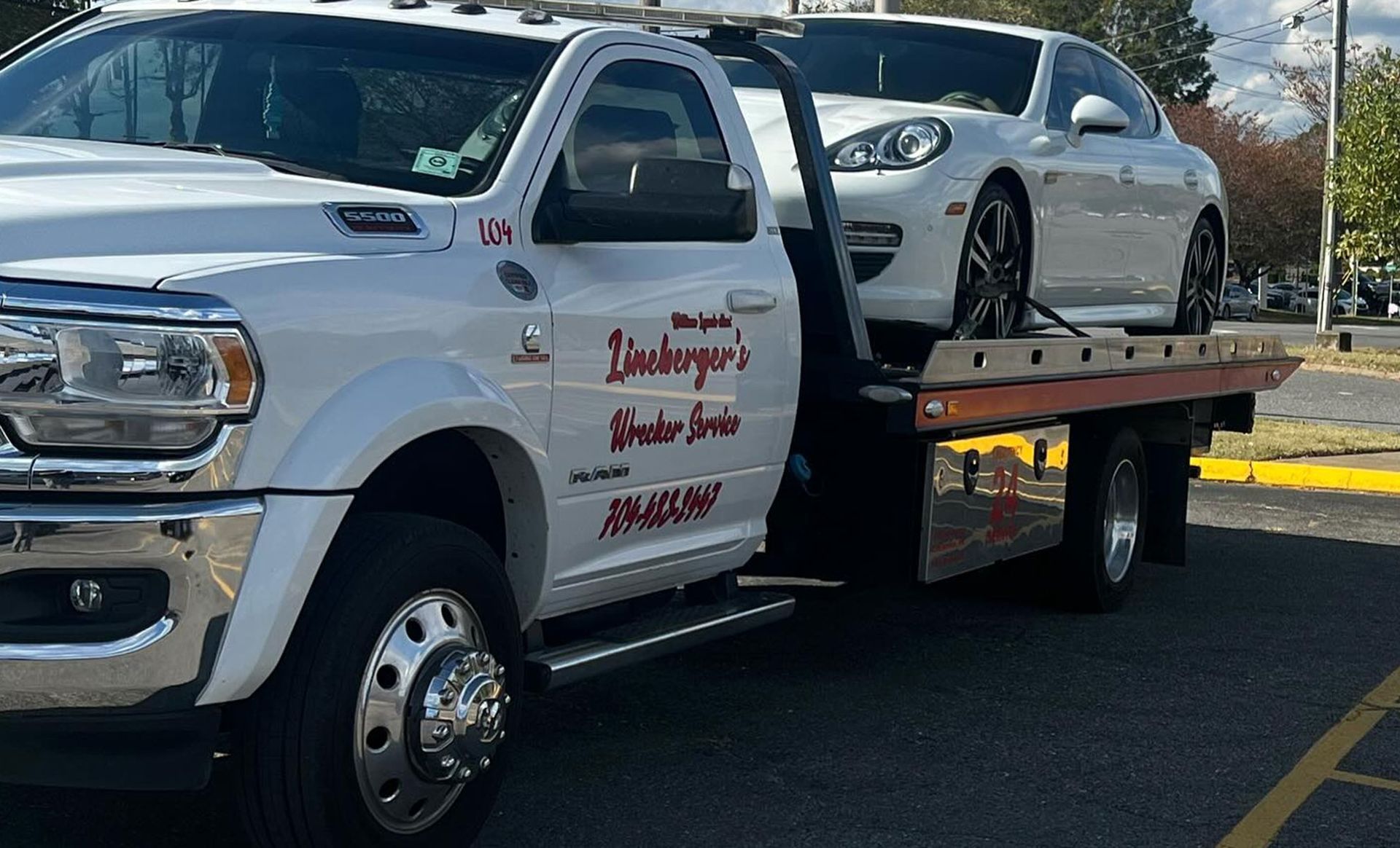 White tow truck carrying a white Porsche on its flatbed. Sunny outdoor setting.