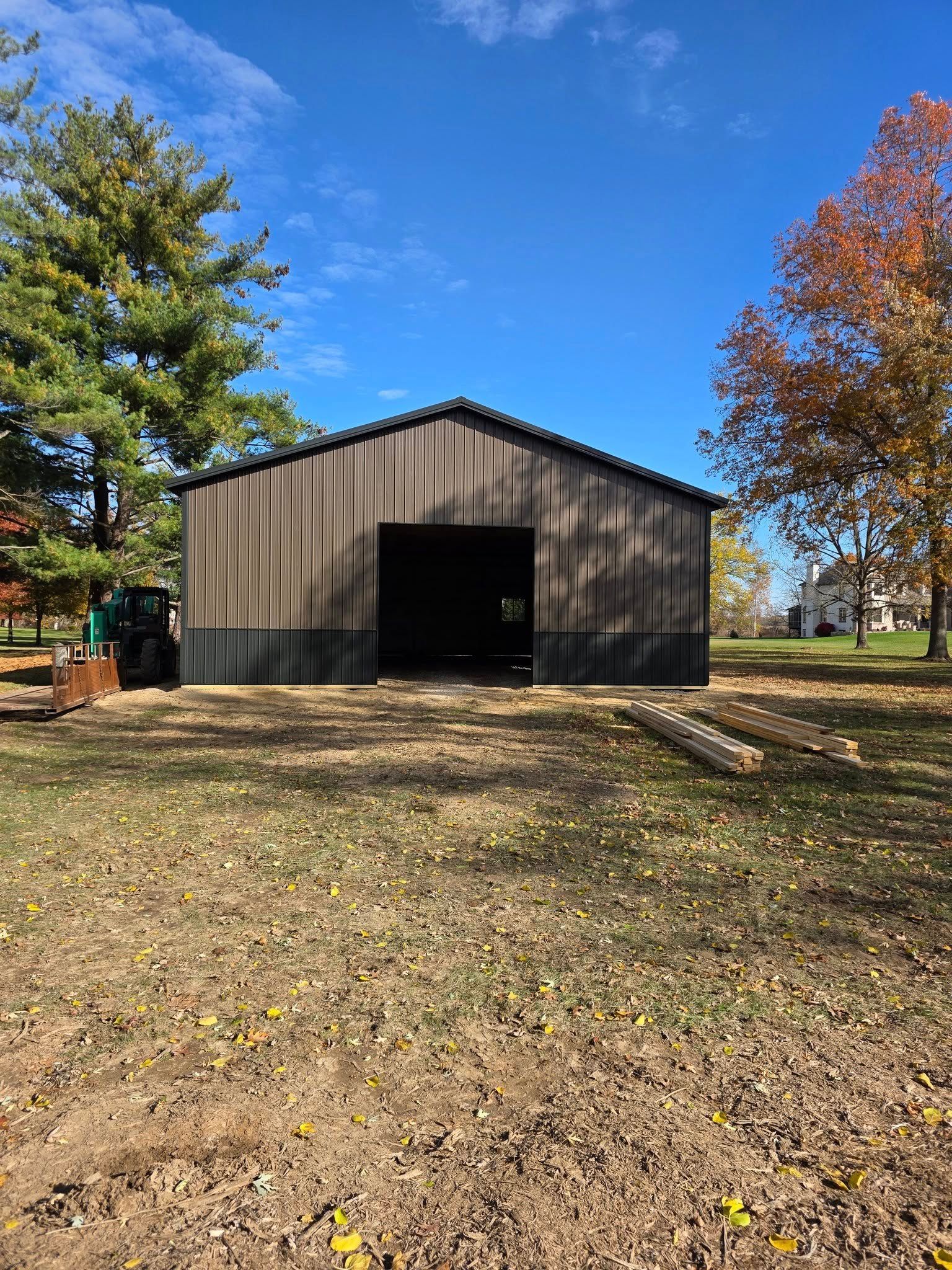 Large brown metal barn with open front on a grassy lawn under a blue sky with trees.