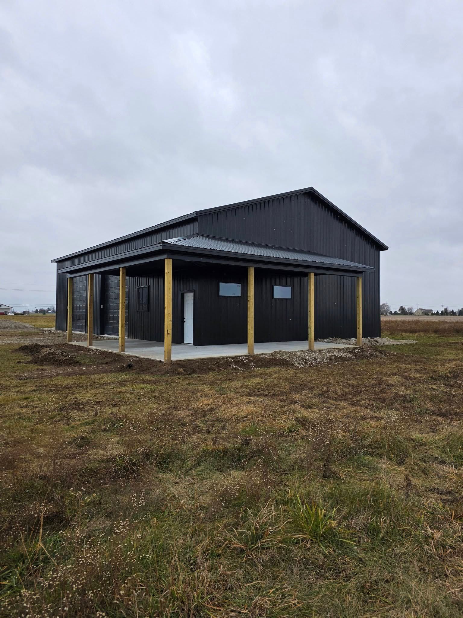 Dark metal-clad building with a porch, set on a concrete pad in a grassy field under an overcast sky.