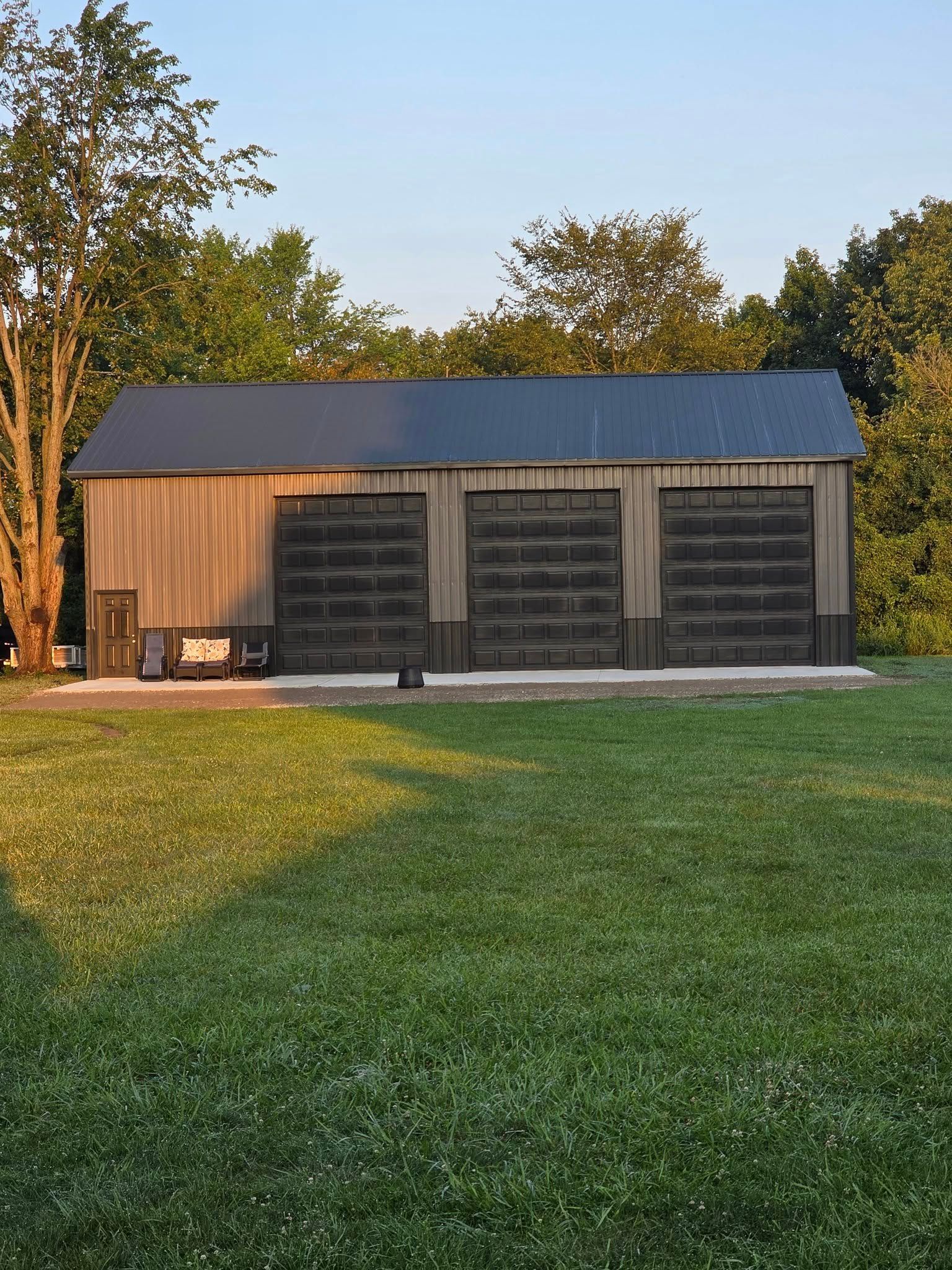 Metal-sided barn with three large garage doors and a dark roof. It's set in a grassy area with trees in the background.