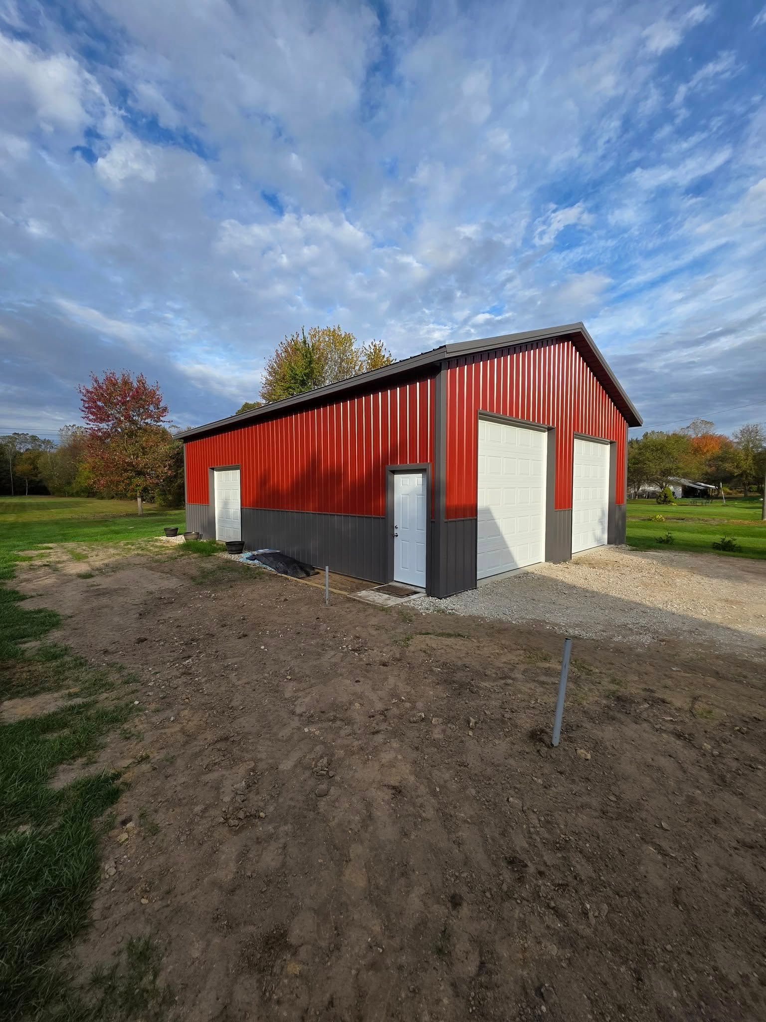 Red and gray barn with white doors on a gravel and dirt lot under a cloudy sky.