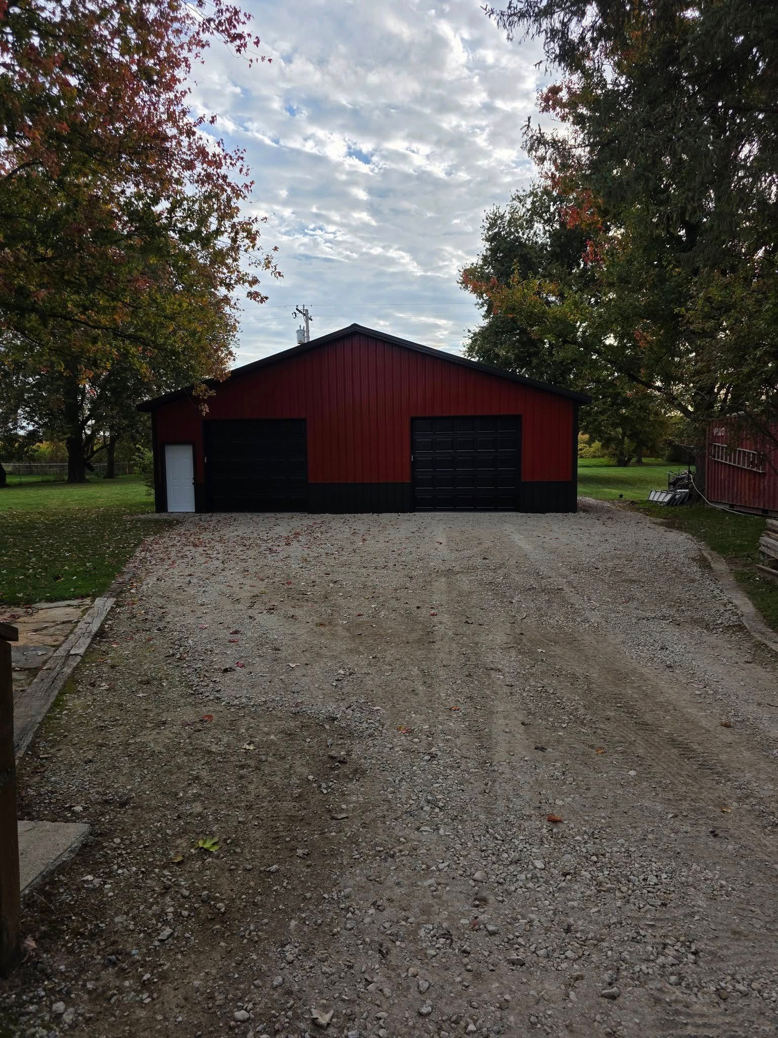 Red garage with black doors and white door, gravel driveway, trees, cloudy sky.