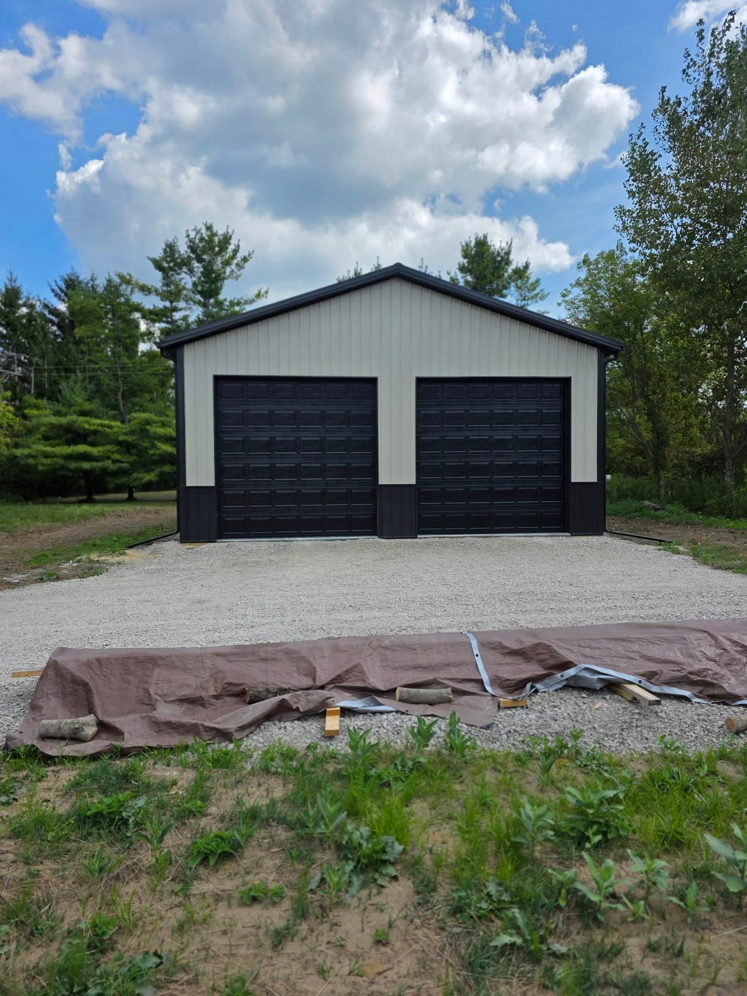 Two-car garage with black doors and trim, beige siding, gravel driveway, and trees under a cloudy sky.