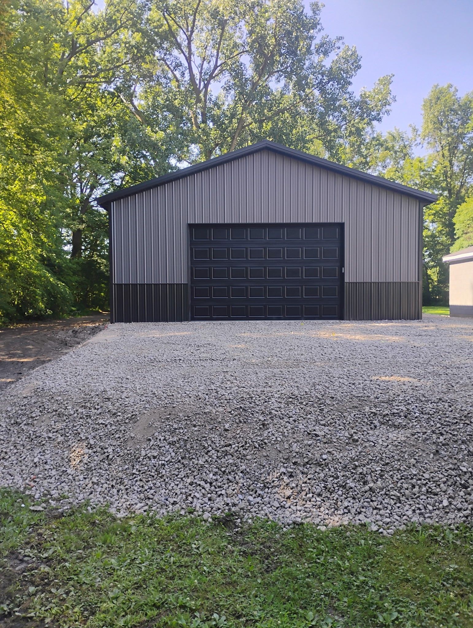 Metal garage with gravel driveway, gray and black siding, black garage door, trees in background.