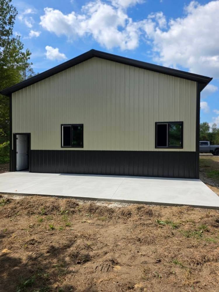 A two-tone metal building with a concrete pad. Exterior features black trim and windows, a tan upper, and a dark base.