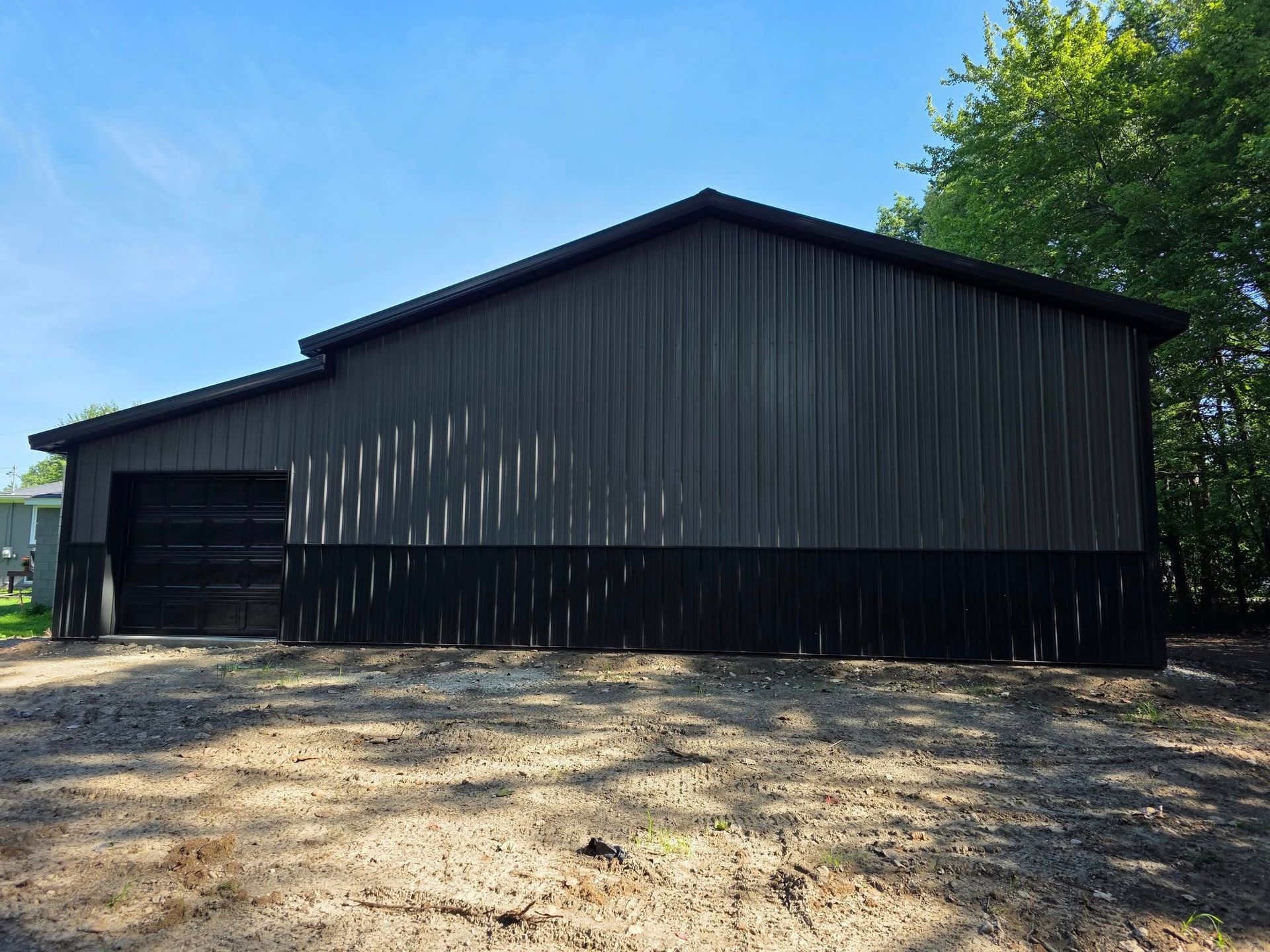 Dark gray metal building with black bottom section and garage door, standing outside with trees and blue sky.