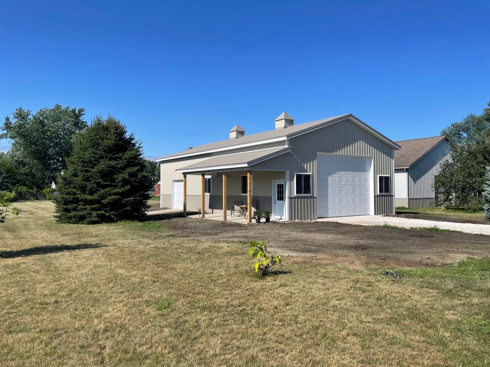 Metal-clad building with a porch, garage door, and small trees on a grassy lot under a blue sky.