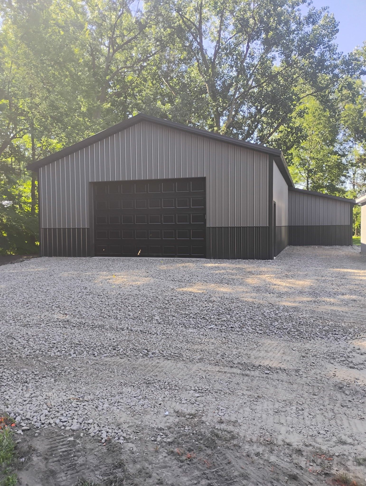 Gray metal barn with black doors and dark gray lower walls, gravel ground, and trees in the background.