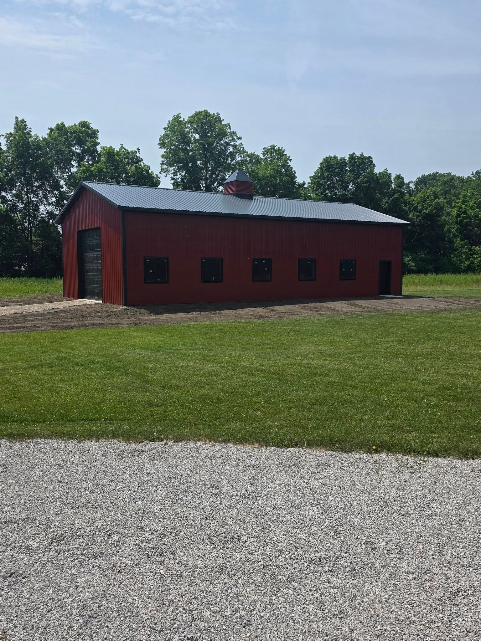 Red barn with dark roof, set on grass field with trees and a gravel path.