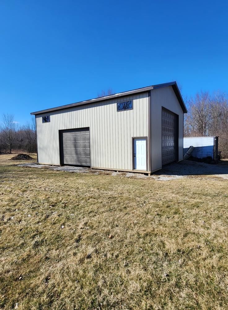 Metal barn with two garage doors and a side door, set on a grassy field under a blue sky.
