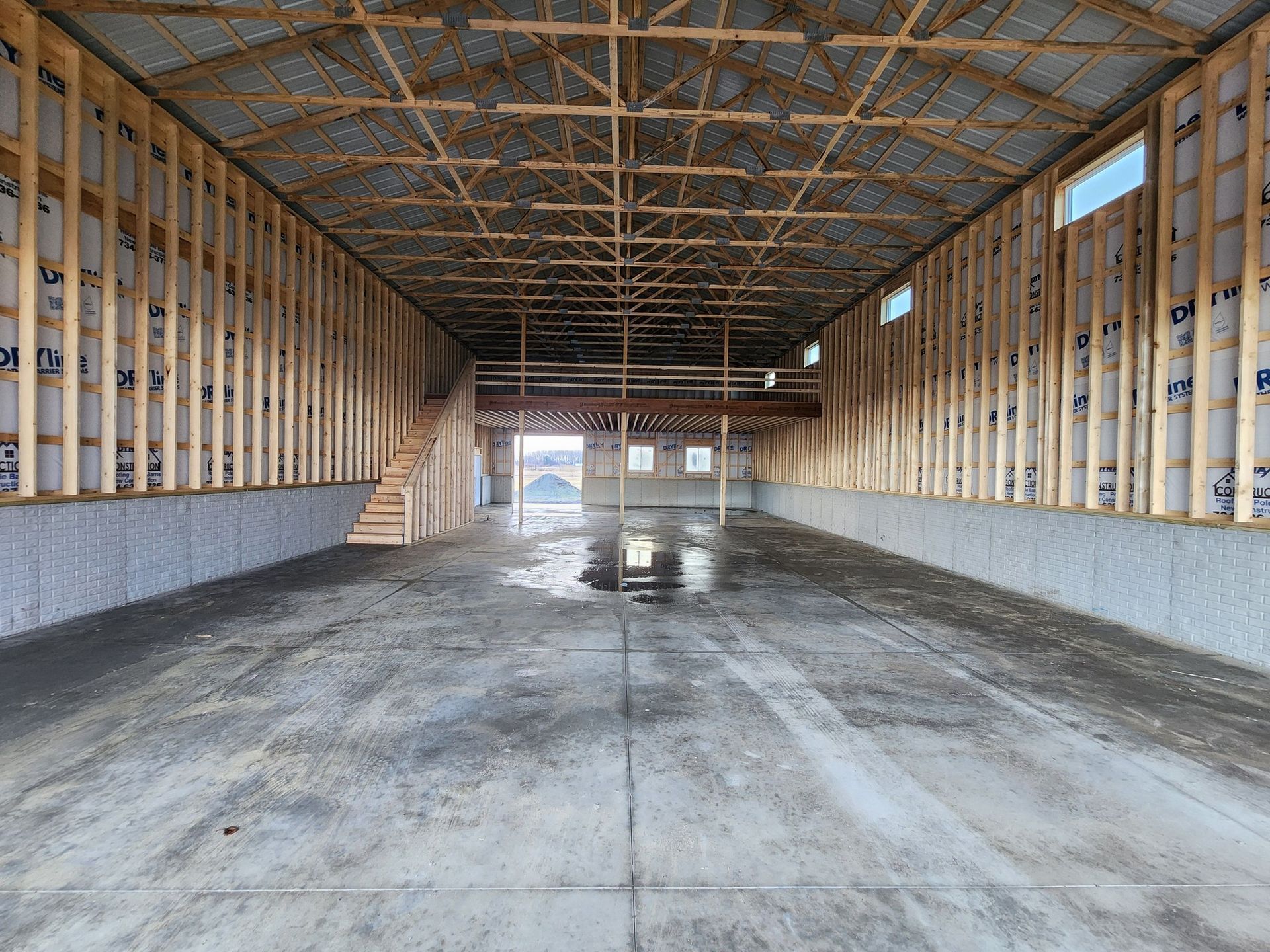 Interior view of a large unfinished building with wooden framework, concrete floor, and small windows.