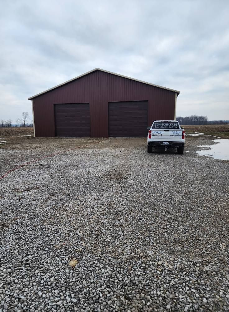 Brown metal barn with two garage doors, parked white truck on gravel. Cloudy sky.