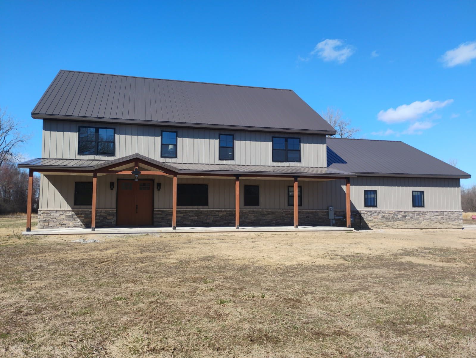 Two-story house with gray metal roof, light brown siding, and stone facade on a sunny day.