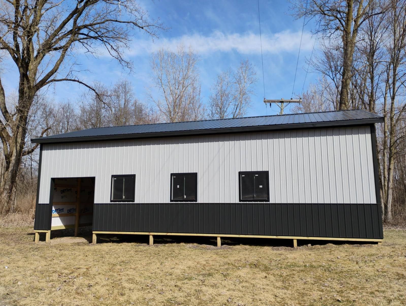Gray and black metal-sided shed with a black roof, three small windows, and open doorway, set against a tree-lined backdrop.