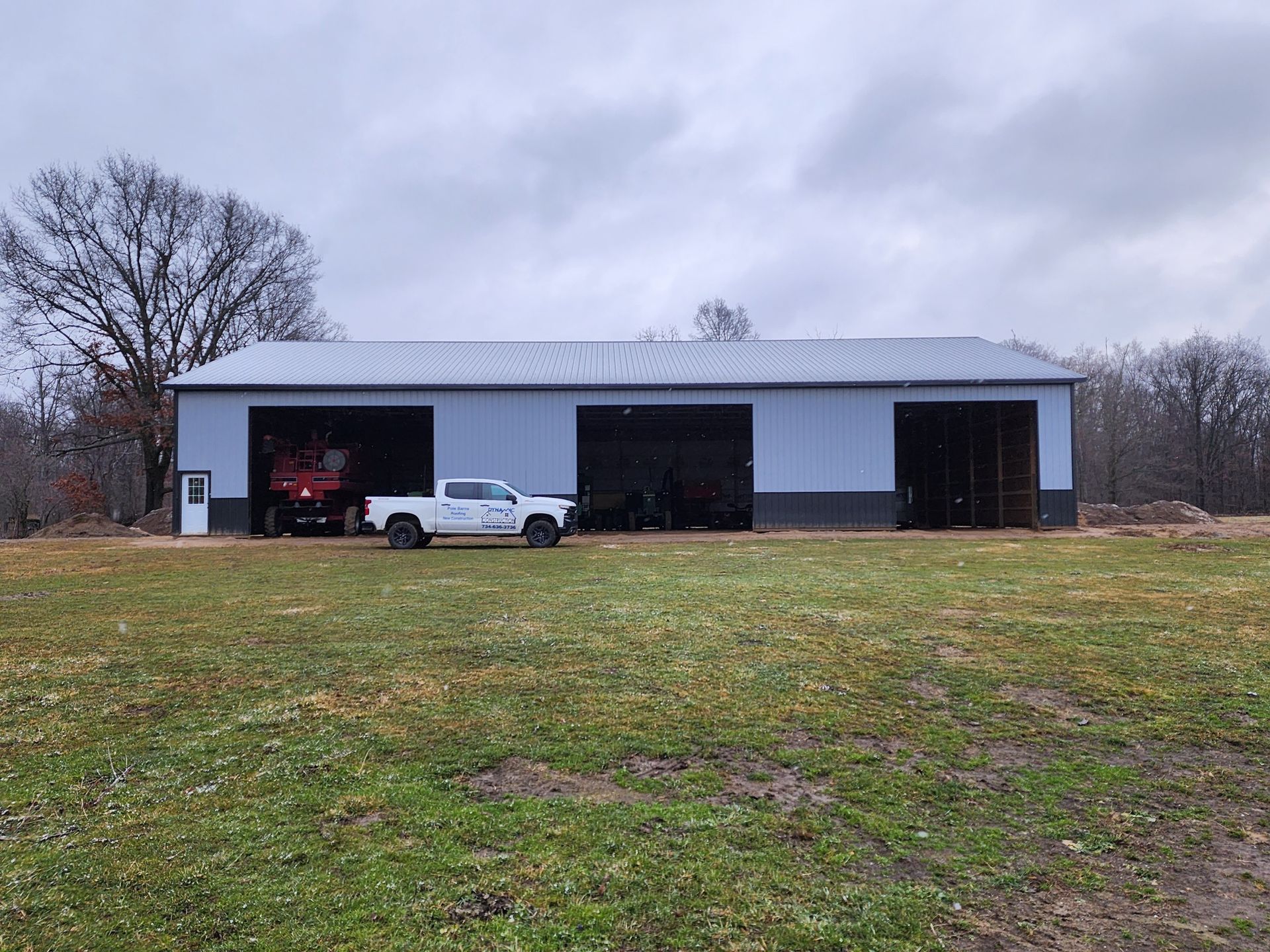 White truck parked in front of a metal barn with three large bays, overcast sky, grassy field.
