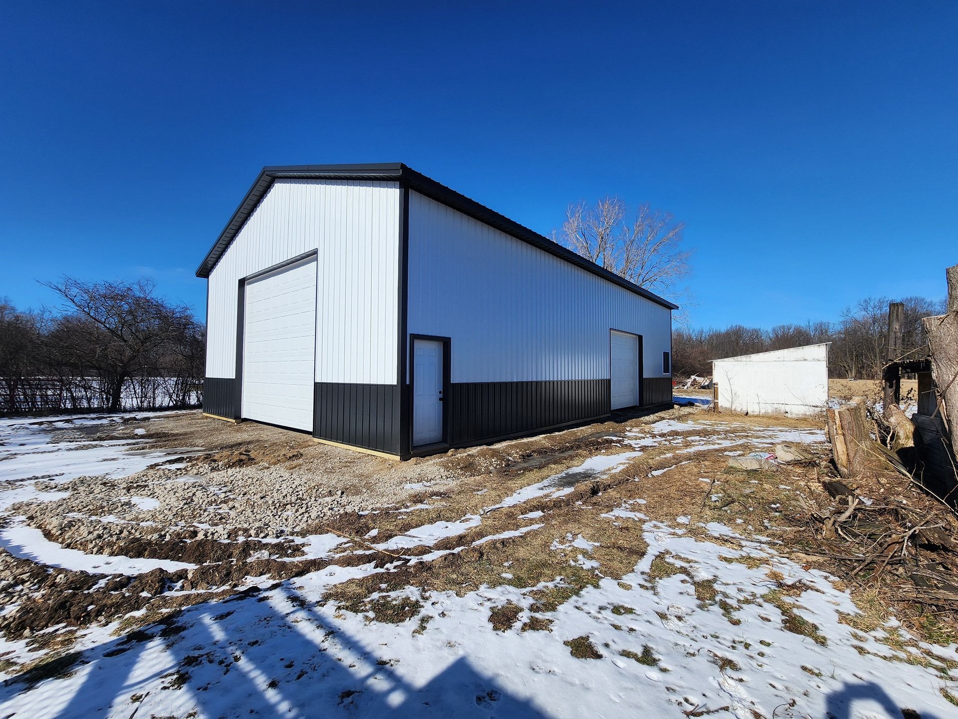 White and black metal barn with roll-up door, small door, and snow-covered ground under a blue sky.