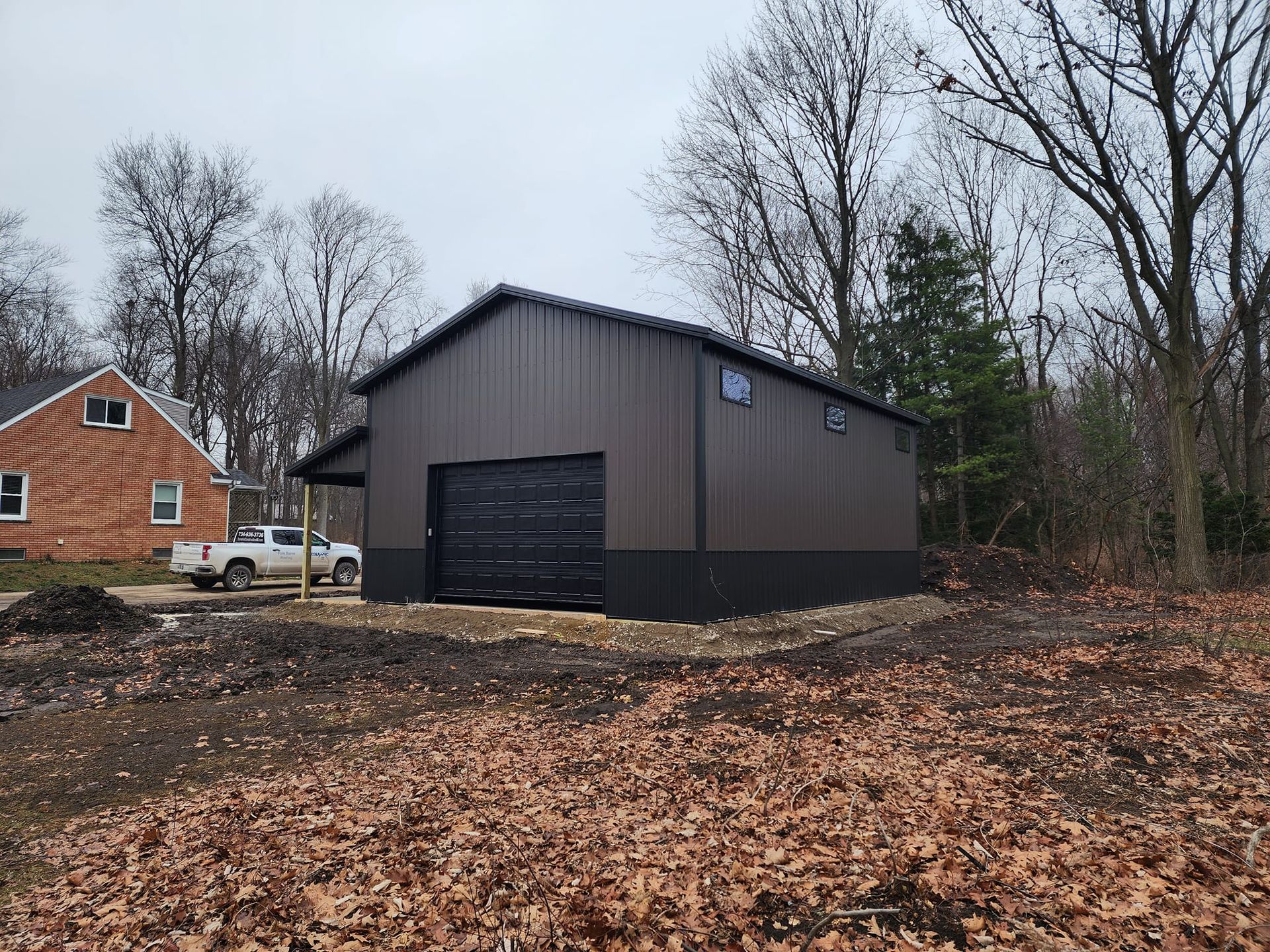 Dark brown metal building with a black garage door and trim, on a lot with bare trees.