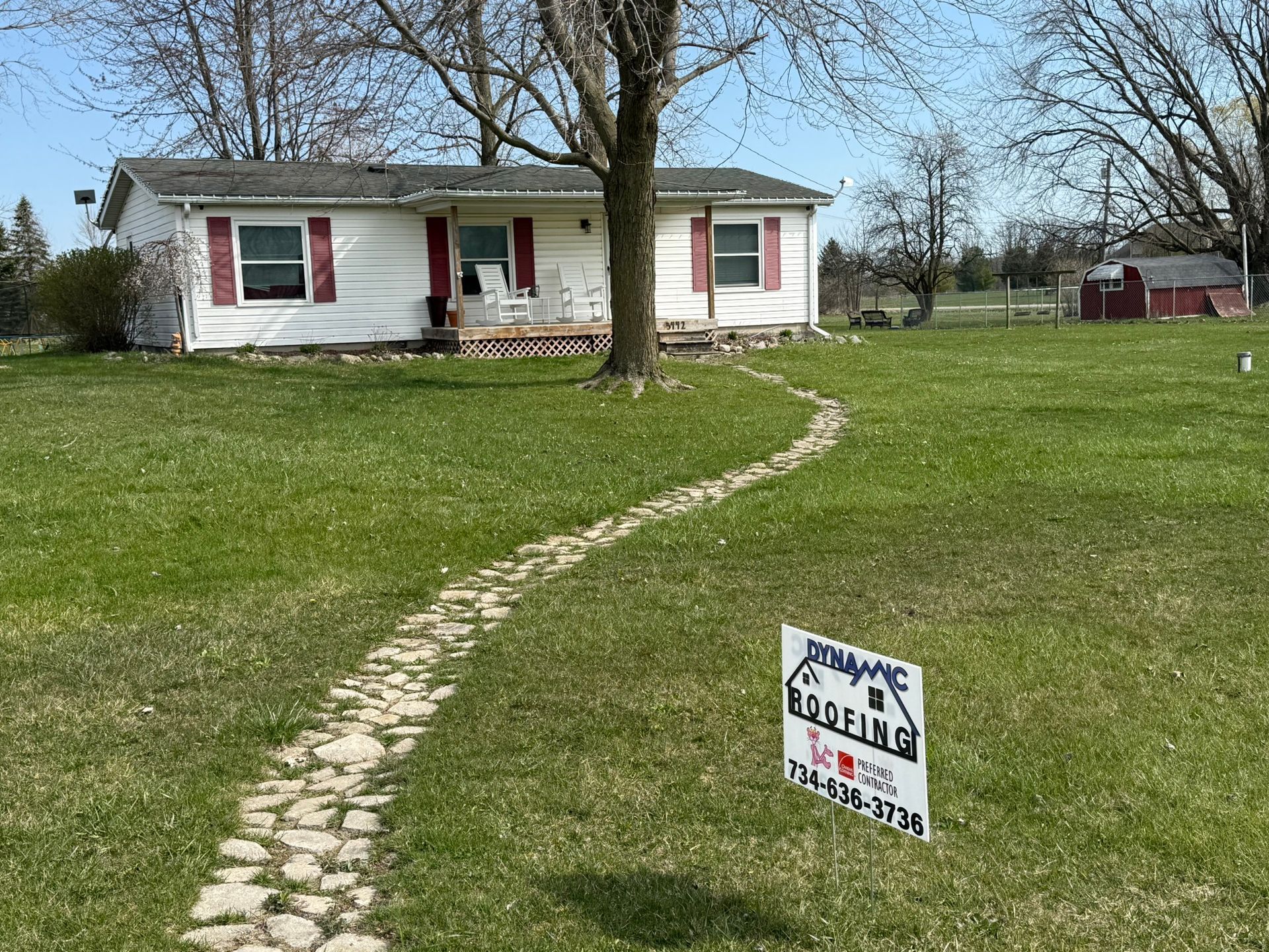 White house with red shutters, a stone path, and a roofing sign in the yard.