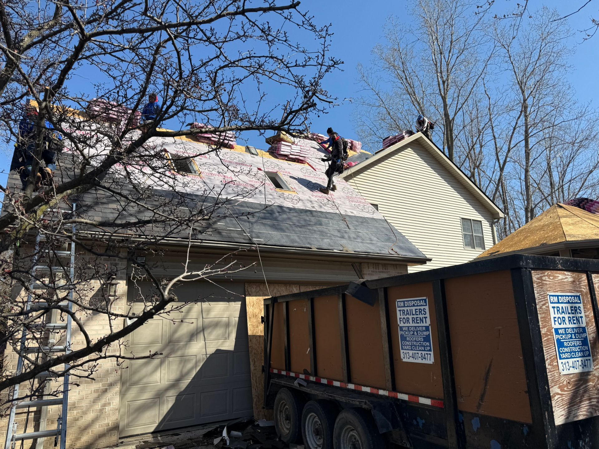 Roofers working on a house, tearing off shingles. Brown trailer in foreground, clear sky.