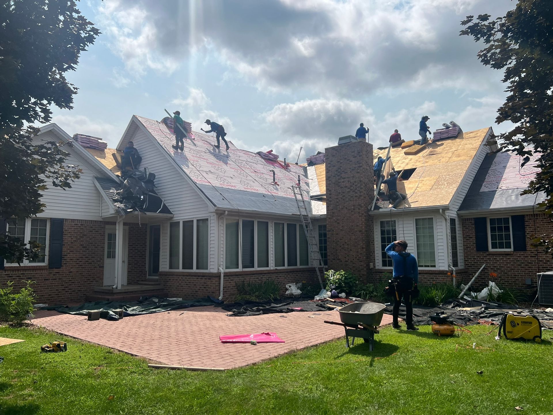 Roofers on a house with exposed wood and brick exterior. Workers wearing safety gear, some on the roof, others below.