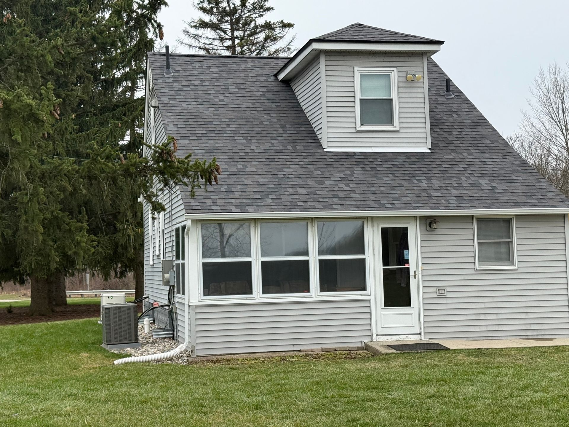 Gray house with a sunroom, dormer, and dark roof, set in a grassy yard.
