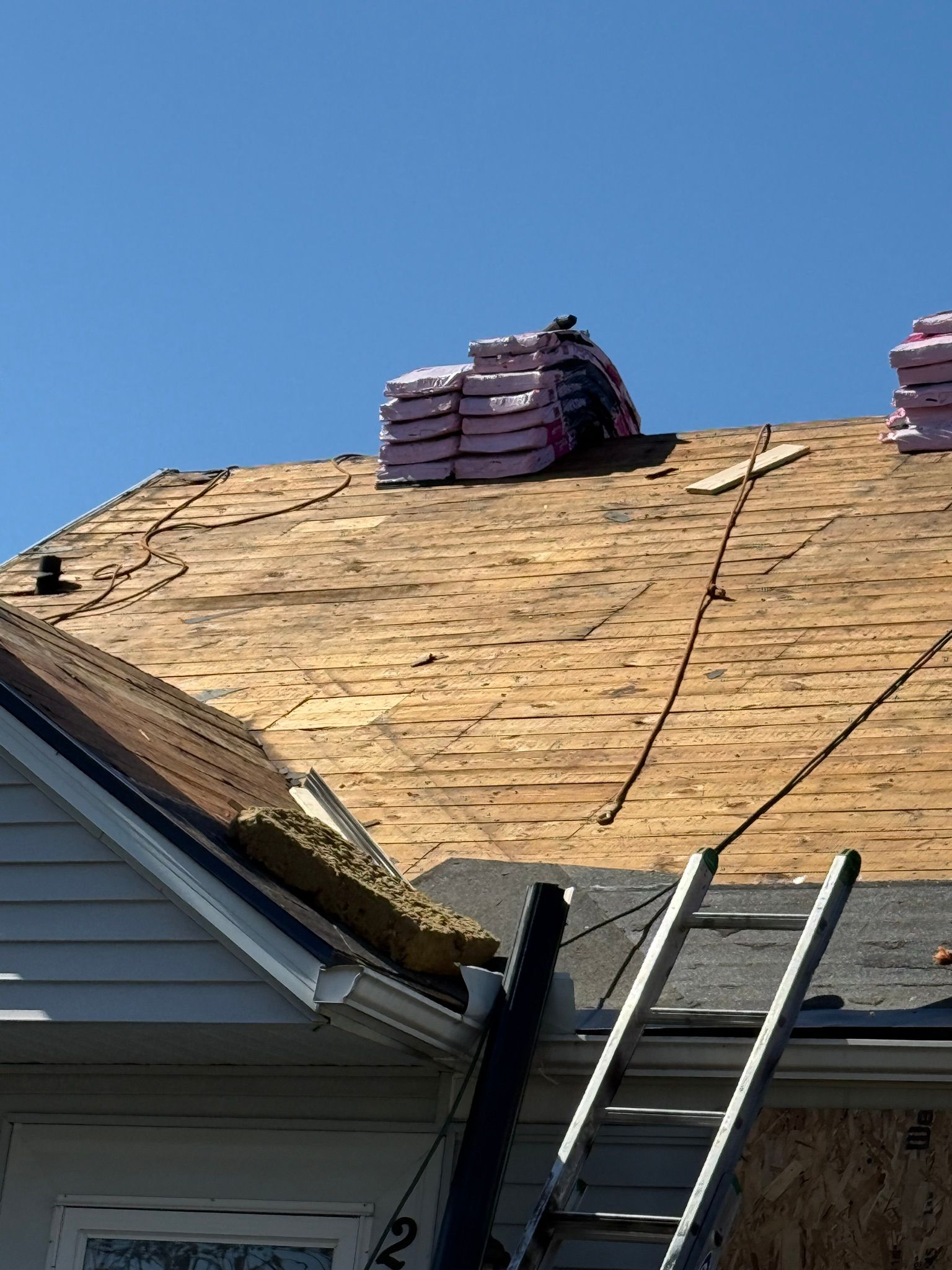 Roof with shingles being replaced; ladder leans against house; blue sky.