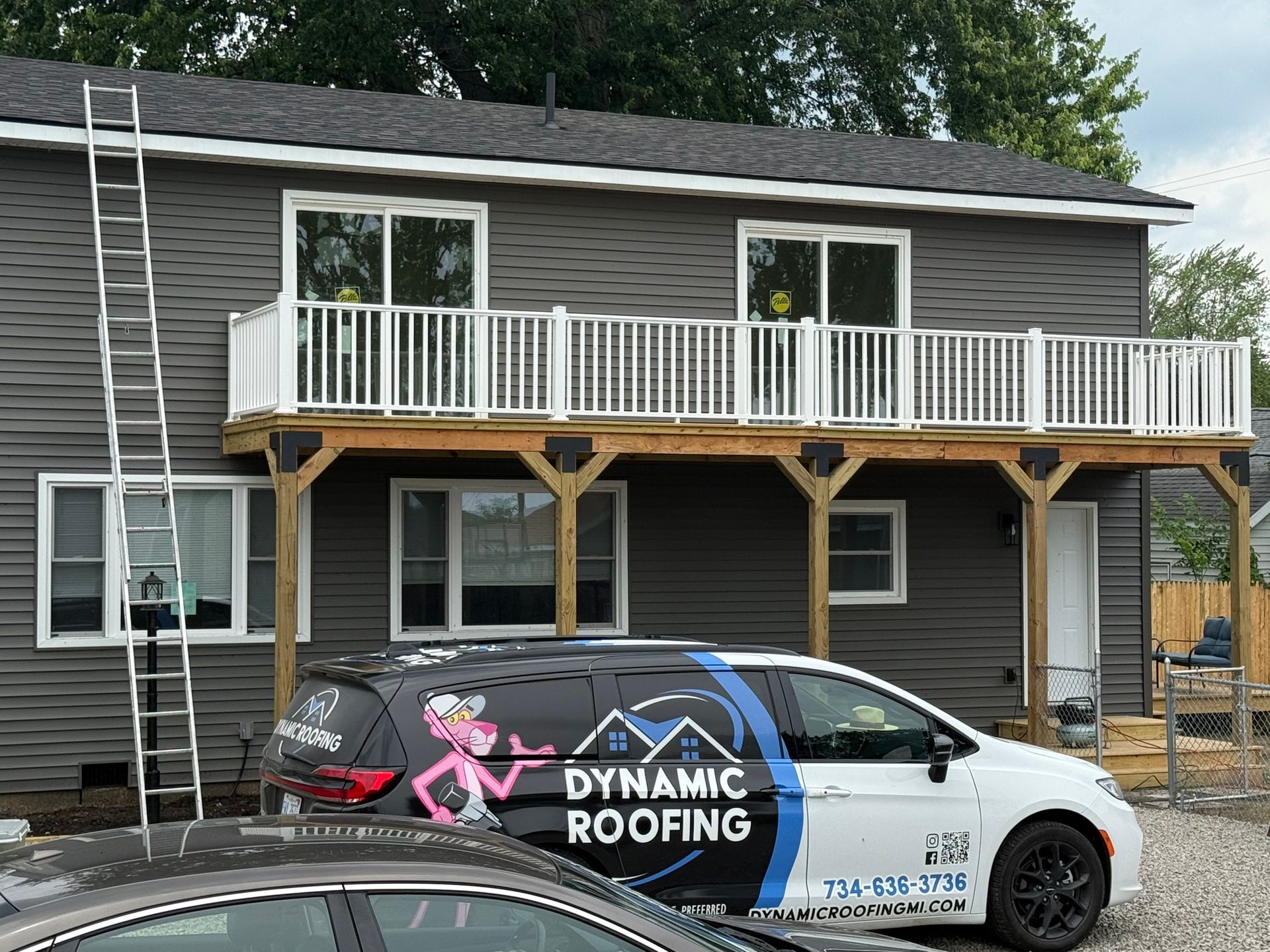 Two-story house with a balcony and Dynamic Roofing van parked in front; ladder propped against the house.