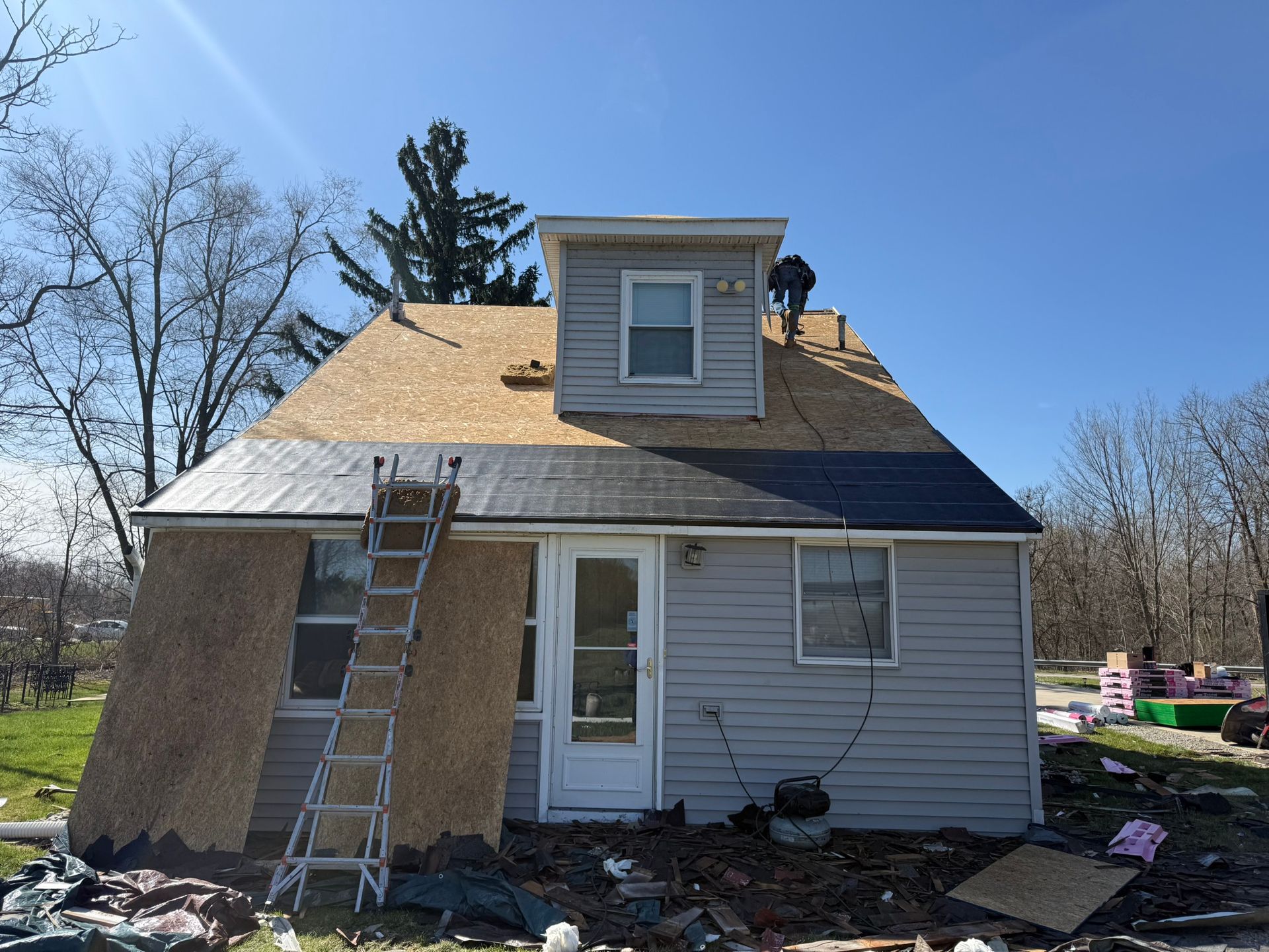 House with partial roof replacement; a worker on the roof, ladder propped against the side, and debris on the ground.