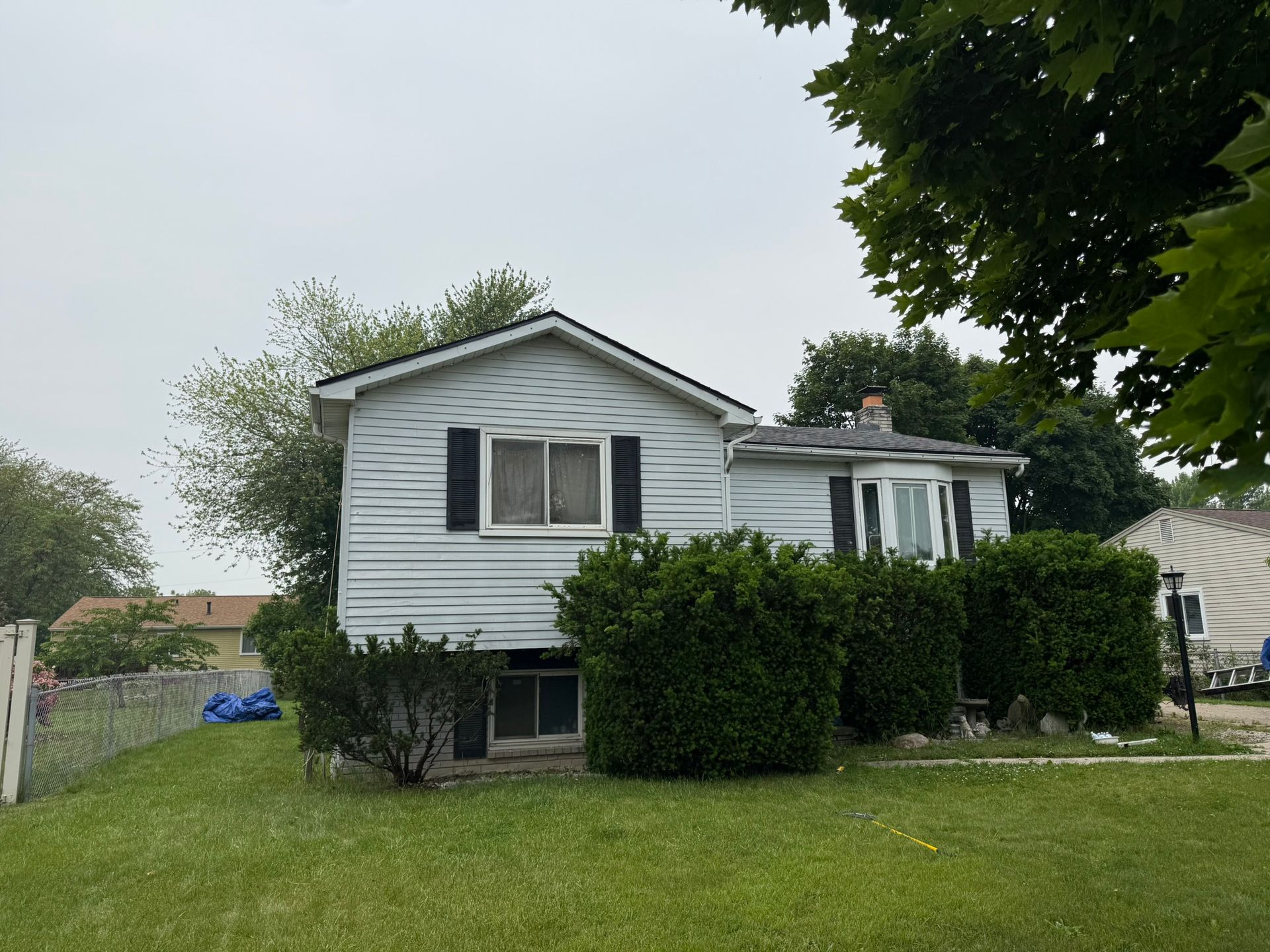 Small, light blue house with black shutters, overgrown bushes, and green lawn under a cloudy sky.