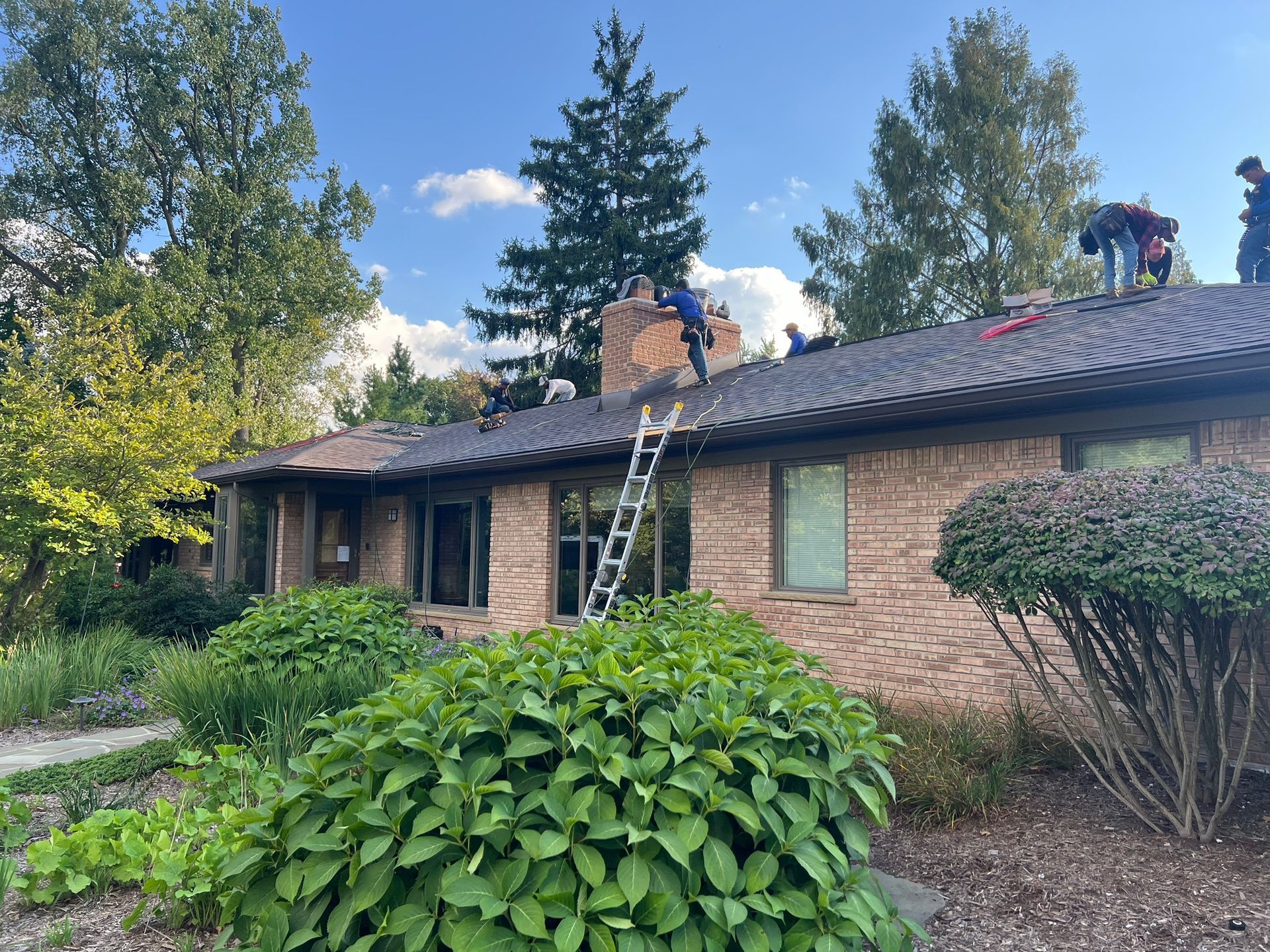 Roofers work on a brick house with trees in the background under a partly cloudy sky.