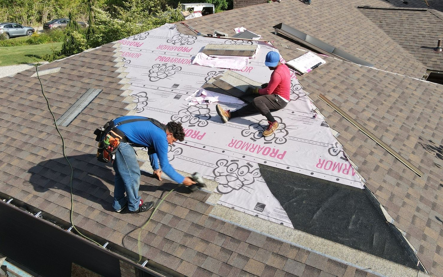 Two roofers replacing shingles on a brown roof. One spreads adhesive, the other sits, holding materials. Sunny day.
