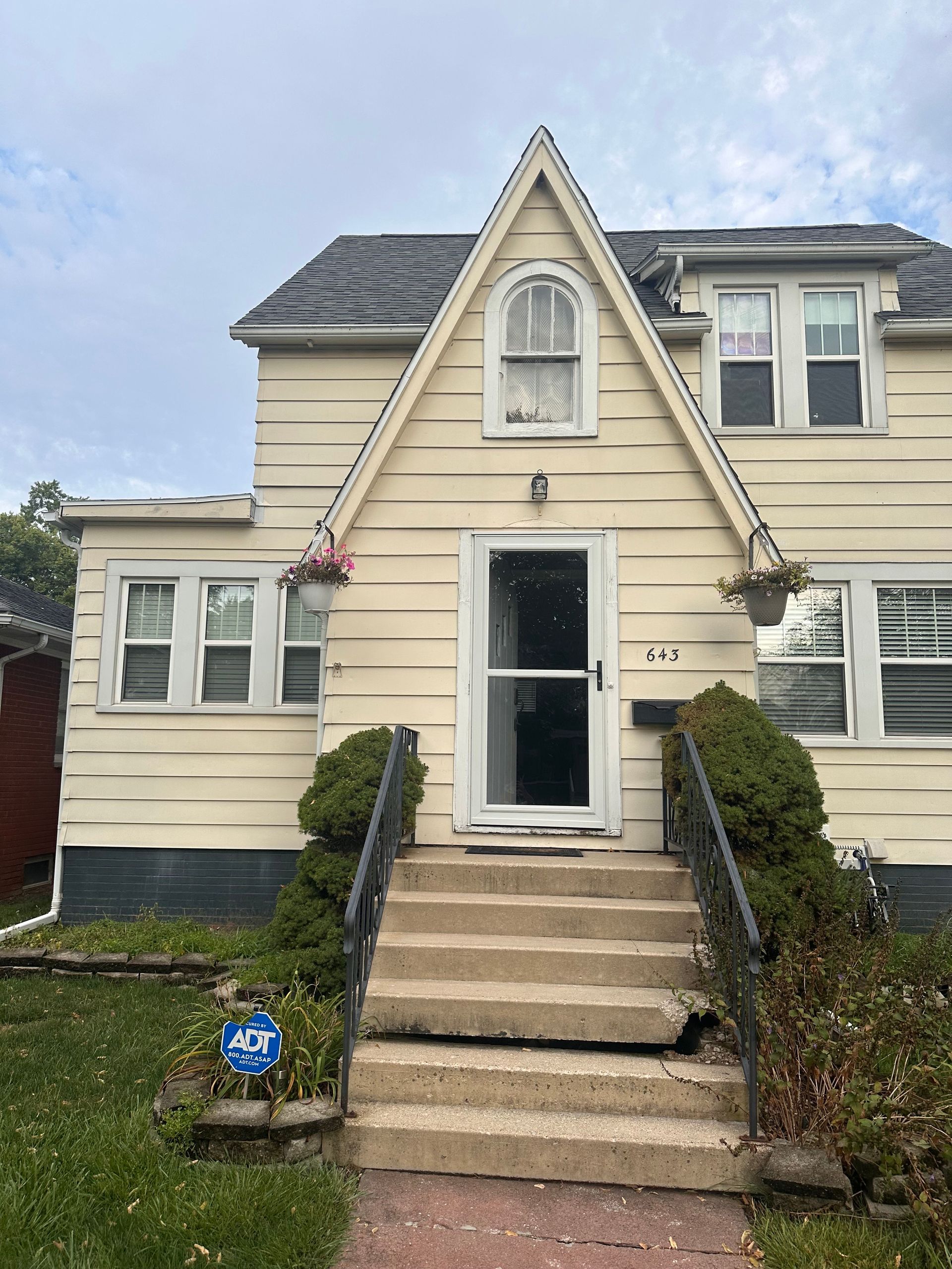 Beige two-story house with a gabled roof, steps, and hanging flower baskets by the entrance.