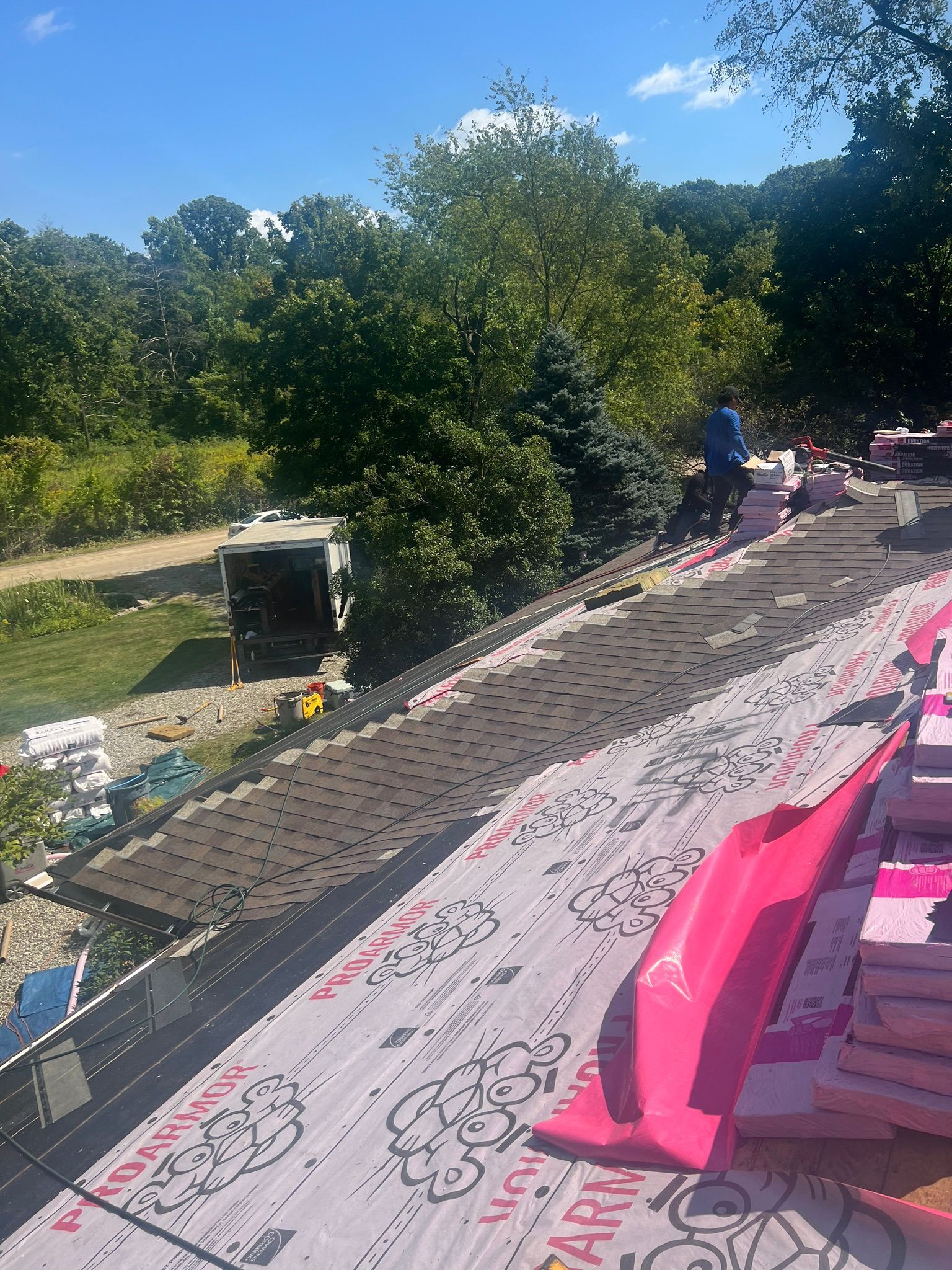 Roofer on a roof, laying shingles. Pink barriers and trees in the background. Sunny day.
