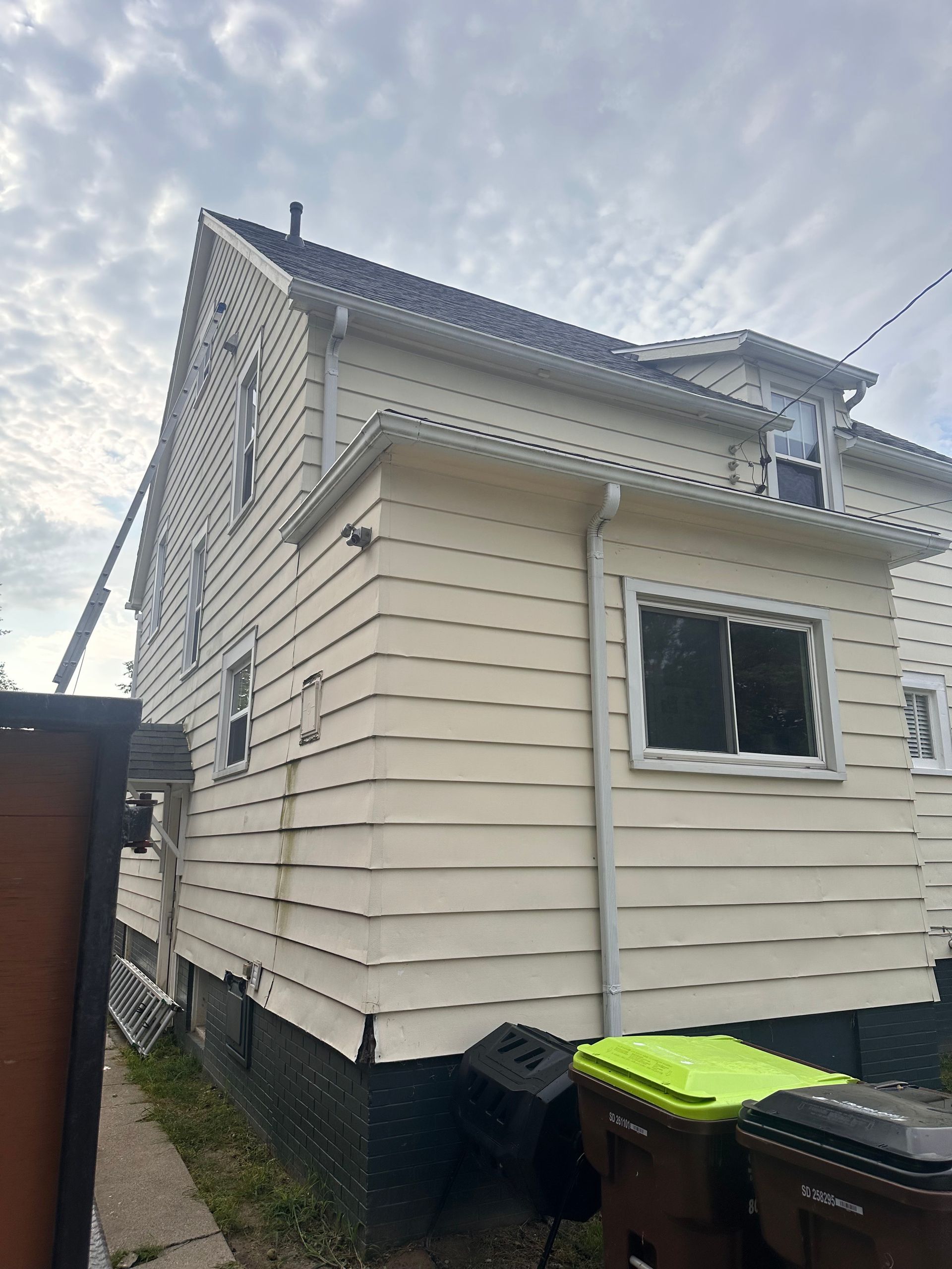 Beige house with white trim, gutters, and windows. Two trash cans sit in front. Overcast sky.